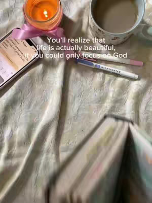 The image shows a cozy and aesthetically pleasing scene. A lit candle sits in a glass holder with a pink ribbon, a cell phone with a screen displaying some content, likely text, a partially visible book, a white mug with what appears to be coffee or tea and a highlighter are arranged on a crumpled, light-colored fabric. The text overlay suggests a message of faith and finding beauty in life.