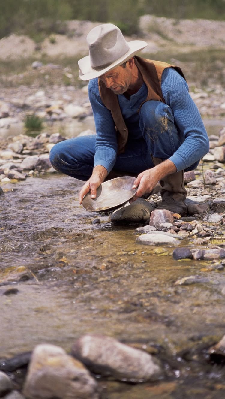 An older, weathered man wearing a cowboy hat, vest, and blue shirt, kneeling in a shallow, rocky riverbed. He is using a pan to sift through the water and rocks, presumably searching for gold. The water is clear, the rocks are gray, and the background suggests a natural, outdoor setting.