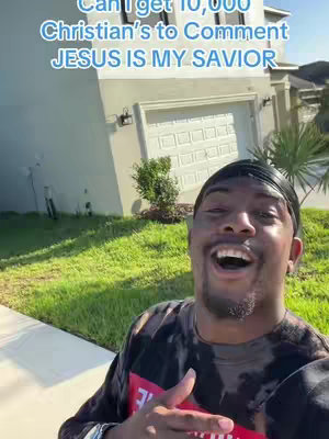 A man with a black durag smiles widely, pointing towards the viewer. In the backdrop, a light-colored house with a white garage door and manicured green lawn establishes the outdoor setting.