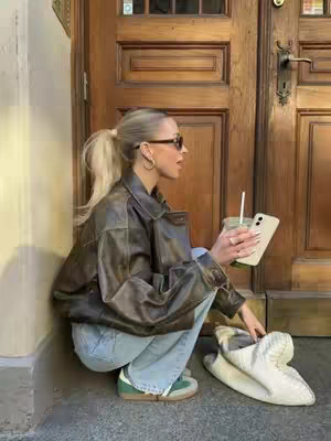A woman with blonde hair in a high ponytail, wearing sunglasses and a brown leather jacket, crouches down on a stoop, holding a phone and a drink with a straw. A pair of white slippers sits beside her next to a wooden door.