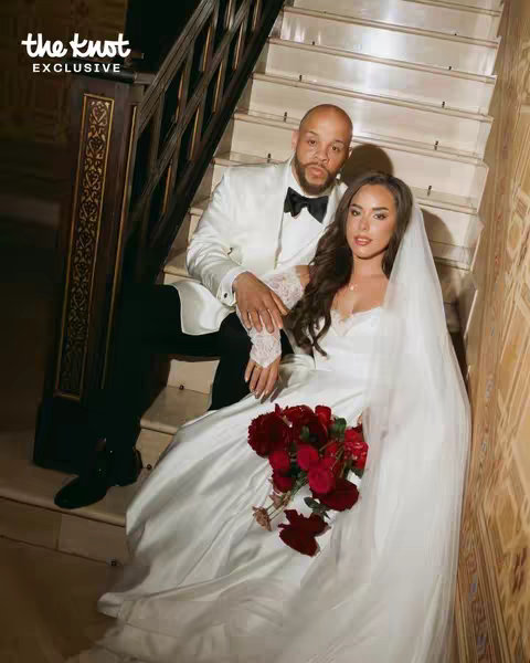 A formal wedding portrait featuring a couple seated on ornate stairs. The woman, in a white wedding dress and veil, holds a bouquet of red roses. The man, in a white tuxedo with a black bow tie, sits beside her. The background includes wood and stone architectural details, suggestive of a luxurious venue.