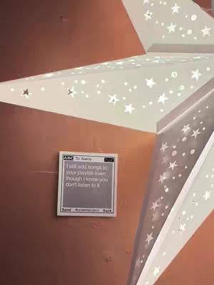 The image shows a star-shaped lamp with cutouts forming star shapes. A small digital note is affixed to the wall, displaying text about adding songs to a playlist. The note is set against a wall with a texture.