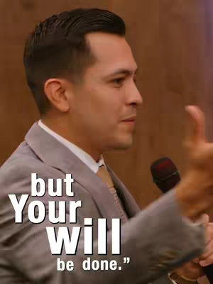 A man in a gray suit speaks into a microphone. The background is blurred and appears to be a room with light-colored wooden panels. Text overlays that read 'but Your Will be done.'
