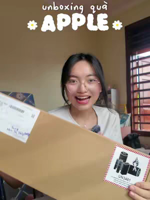 A young woman with glasses smiles widely, holding a large package, likely an Apple product. The background is a domestic setting; the image includes text overlay.