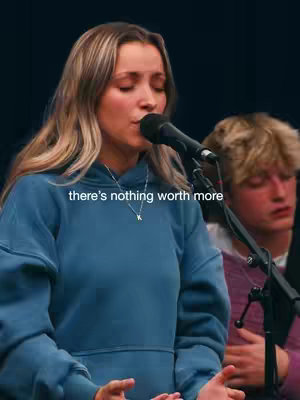 A young woman with long, blonde hair is singing into a microphone. She is wearing a blue hoodie and a silver necklace with a pendant. The background is somewhat blurred, but it appears to be a performance setting with another person partially visible behind her. Text overlayed on the image says, "there's nothing worth more.