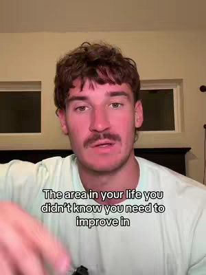 A man in a light green shirt is speaking directly to the camera. Text overlay at the bottom of the screen reads "The area in your life you didn't know you need to improve in". The background is slightly blurred, revealing a bedroom interior with cream-colored walls and two small windows.