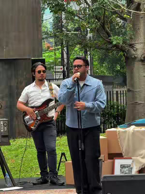 A cover photo captures a man singing into a microphone while another man plays a bass guitar. They stand on a stage outdoors. The singer, in a blue jacket, holds a microphone, while the guitarist is in a white shirt. The scene is framed by out-of-focus greenery and a sign in the background.