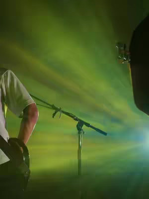 The image shows a portion of a guitarist from the chest up with a microphone stand and stage lighting in the background. The guitarist is wearing a white t-shirt and has his right arm visible, holding the guitar. The lighting creates green and yellow streaks across the background. The top right has a glimpse of a guitar