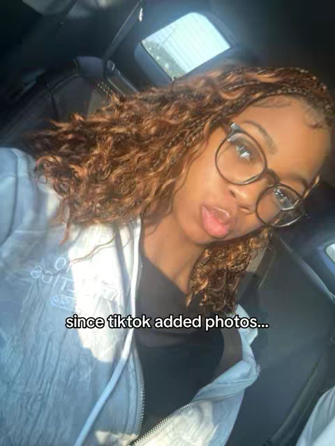 A selfie-style image featuring a young woman in a car, gazing towards the camera. She has long, curly brown hair, large black-framed glasses, and is wearing a white casual top. The interior of the car, including the seat and a window with outside light, forms the backdrop. A text overlay states, 'since tiktok added photos...'