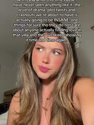 A woman with long, light brown hair is centered in the frame. She has fair skin, a slight smile, and is looking slightly off-center, as if listening or thinking. The background is blurred, suggesting a casual setting. The image has a warm color palette, with natural lighting.