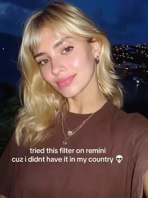 A selfie-style shot taken at night with a woman in the foreground. She has long, blonde hair with bangs, and is wearing a brown top. The background shows city lights under a dark, possibly twilight, sky. The woman is looking at the camera with her lips slightly parted.