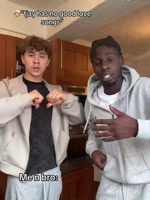 Two young men, one with curly light brown hair and a light colored zip-up jacket, stands on the left, making hand gestures. Another ebony-skinned man in a light hoodie is on the right, gesturing as if talking. The background shows wood-paneled cabinets, suggesting an indoor setting.