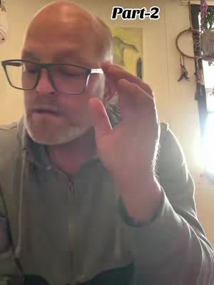 A middle-aged man with glasses is the central focus, seen from roughly the chest up. He wears glasses and a grey zip-up, and appears to be in mid-sentence or expression. The backdrop includes a wall with an abstract painting and a window, softly lit.