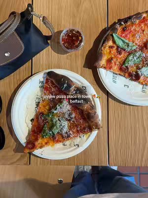 Overhead shot of a wooden table with food. There's a plate with a slice of pizza and another half-pizza slice on a different plate. A small, clear container with a red sauce. A black purse and a brown strap are next to the pizza. The image uses warm lighting and a bright, inviting composition.