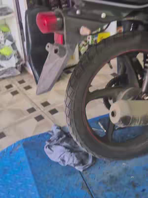 The image shows the rear wheel and part of the suspension of a motorcycle. The tire is black with a red stripe around the rim. The surroundings feature a blue surface and tiled floor.
