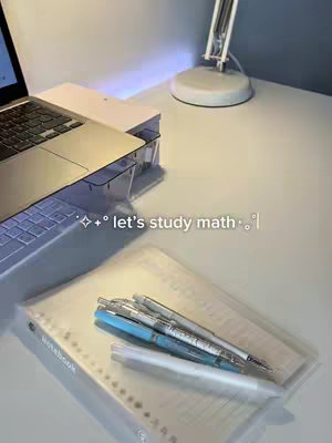 A well-lit desk arrangement with a laptop, a clear plastic pen holder, and a white desk lamp. Pens and a notebook are placed on the desk surface. A laptop is displayed in the upper left corner, and the desk lamp is in the upper right.