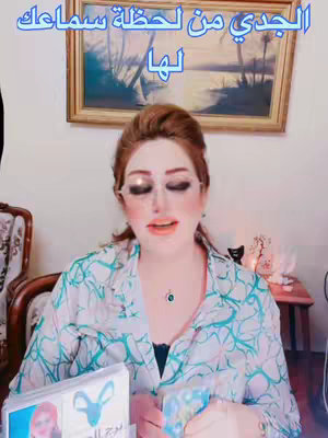A woman with reddish-brown hair and makeup sits in front of the camera. She's holding tarot cards and looking forward. Behind her is a painting in a wooden frame.