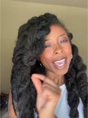 A woman with long, dark braided hair is shown in a selfie-style video. She is wearing a white top and has a nose ring. She is speaking with her mouth open, and her hand is raised near her face with her fingers pointing outwards.