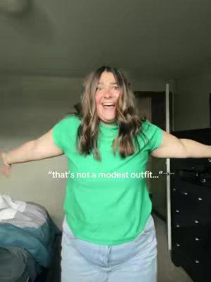 A woman with long wavy hair stands in a room with her arms outstretched, smiling and laughing with an excited expression. She's wearing a green t-shirt and light blue jeans. The background is blurred, showing a glimpse of a bed and a dresser. A text overlay reads, "that's not a modest outfit..."