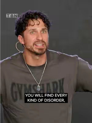 The cover image features a man with short, curly dark hair, wearing a gray Gymshark t-shirt and a silver necklace. He has a microphone clipped to his ear. Overlaid on the bottom is a black rectangle with white text.