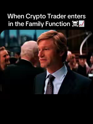 A still from a movie featuring a man in a suit, with text overlay at the top. The man is the central focus, with a crowd behind him, and the background is blurred. The man has short, light brown hair, and is wearing a dark suit, white shirt, and a patterned tie. The lighting appears to be a mix of indoor and natural light.