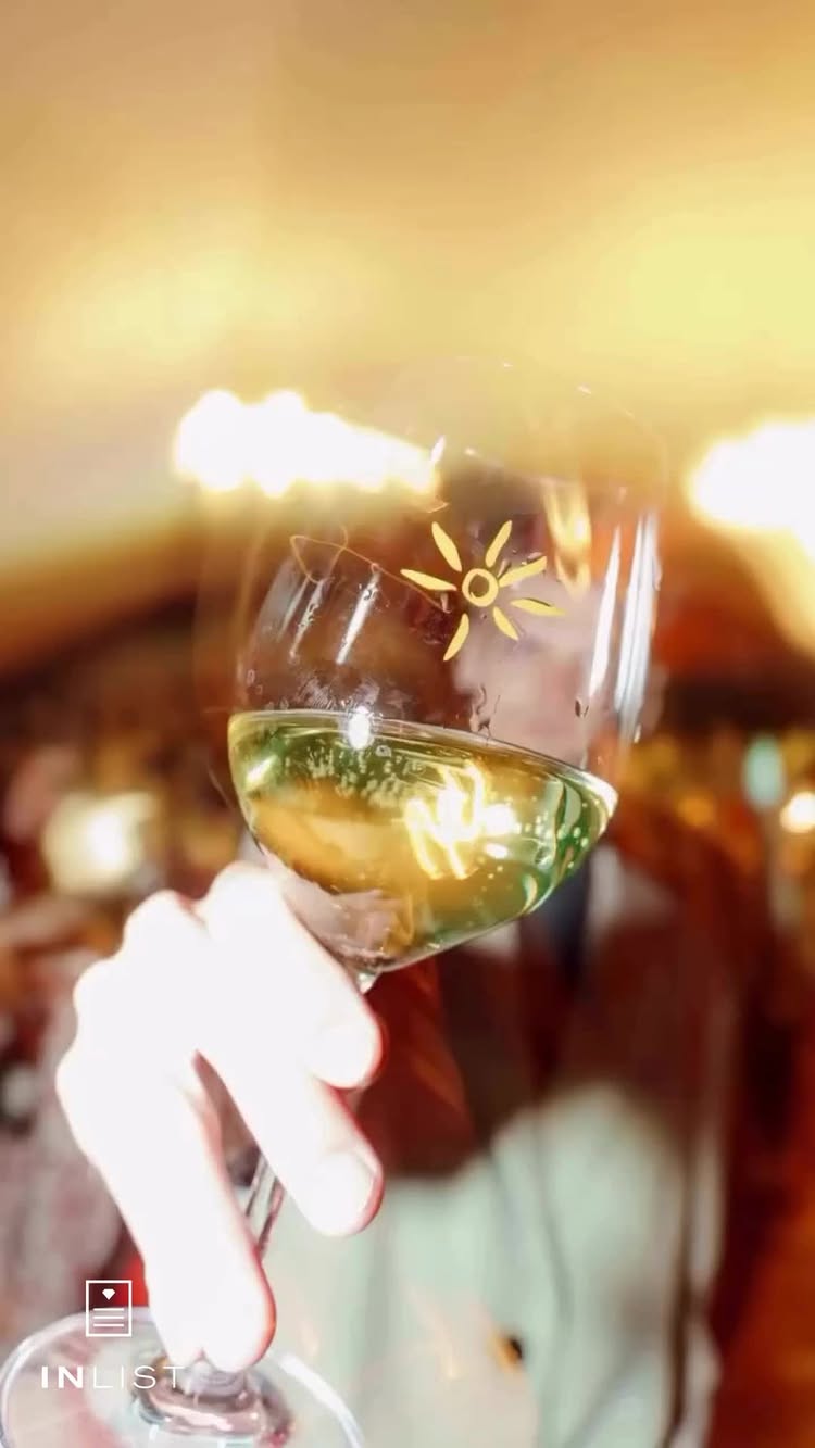 The image shows a close-up view of a hand holding a wine glass, which is filled with a light-colored liquid, likely wine. The glass has a decorative element of a sun design on it. The background is blurred, indicating an indoor restaurant setting. A logo is present at the bottom left corner. The overall composition gives off an air of sophistication.