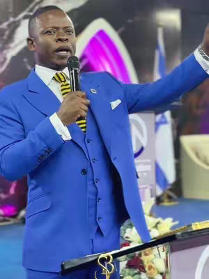 A man in a blue suit, standing in front of a podium, speaking into a microphone with one arm raised. The background includes a church architecture.