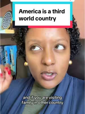 A selfie-style shot featuring a woman of color with curly hair, wearing a blue top and gold hoop earrings. A text overlay in a block above her head reads, "America is a third world country". The background suggests an office or home setting, with a shelf and some items visible. The woman is looking upwards, possibly at the text, and speaking.