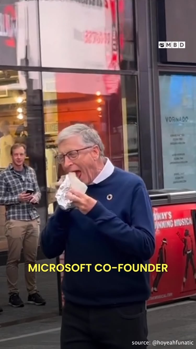 Bill Gates eating a wrap outside in front of a store with text overlay at the bottom of the screen.
