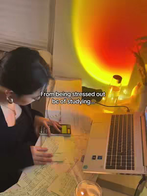 A person with black hair and glasses is studying at a desk. The desk is cluttered with papers, a calculator, and a laptop. A lamp casts a warm yellow glow on the scene. The person appears to be focused on their study materials.