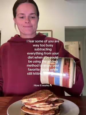 A woman in a burgundy hoodie is captured from the chest up, slightly angled to the side, smiling. She's holding a colorful mug, appearing to pour something onto a plate of pancakes. The pancakes are stacked on a white plate sitting atop a dark wooden table. Behind her, a framed picture hangs on the wall.