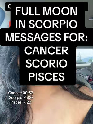A selfie-style shot featuring a woman's upper body and face. The focal point is a black rectangular text overlay that dominates the image. The text itself, in a bold, white sans-serif font, announces "FULL MOON IN SCORPIO MESSAGES FOR:", followed by a list of zodiac signs: CANCER, SCORPIO, and PISCES. Below the astrological signs, the video includes timestamps for each sign's segment. The woman's dark hair is visible, and partial views of a car interior with light blue-grey seats and window view in the background. The photo is taken in daylight.