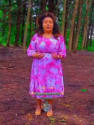 A woman, wearing a pink dress, stands in a forest setting. The dress features a floral pattern. Trees are visible in the background.