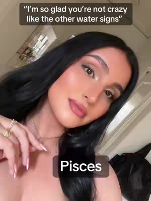 A woman with long black hair and makeup gazes slightly off-center. Above her, a text overlay says, "I'm so glad you're not crazy like the other water signs". At the bottom, a brown box says "Pisces". The background suggests a well-lit indoor area with soft lighting.