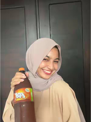 A woman in a beige hijab and shirt smiles broadly while holding a large bottle of beverage, likely soda. The background is a dark wooden door.