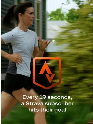 A woman in athletic attire is running outdoors. The image features a blurry, green background hinting at trees and foliage, with a road or path visible. A large, orange Strava logo is prominently displayed. White text overlays the image, promoting the app.