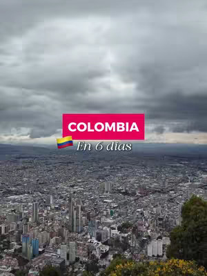 An aerial view of a city under a cloudy sky. A red rectangular banner displays 'COLOMBIA' and 'En 6 días' in white text with the Colombian flag in the bottom left corner. There are trees in the foreground and a large city in the background with skyscrapers.