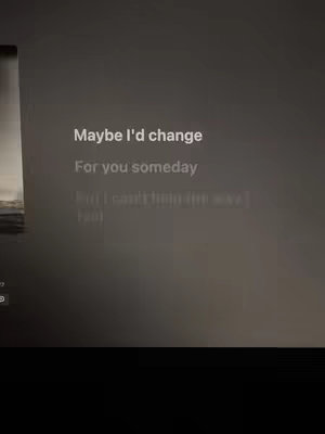 The image shows a dark screen displaying song lyrics. The text 'Maybe I'd change For you someday' is prominently displayed in a clean, sans-serif font. Beneath it, a continuation of the lyrics is partially visible. The background appears to be a blurred image, and the overall feel is simple and focused on the text.