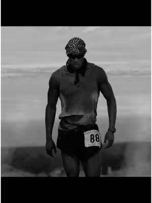 A black and white image of a muscular man running. He wears a tank top, shorts and a bandana. His shirt is ripped and his face is covered with sweat. He has a race number pinned to his shorts. The background appears to be a cloudy sky.