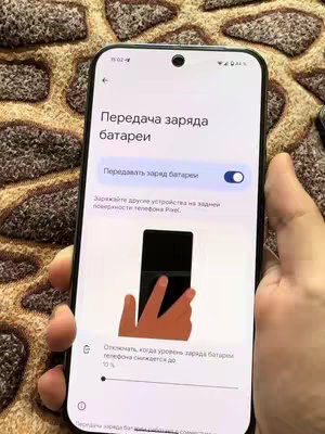 A smartphone screen is held in a hand, displaying a settings menu with Russian text, a graphic representing battery charging, and the Pixel phone logo. The background is textured with brown and tan patterns.