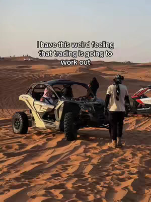 The image shows an off-road vehicle in a desert environment. The foreground is dominated by sand dunes, with the vehicle positioned on top of them. A person appears to be walking toward the vehicle on the right. In the middle ground, the vehicle, a buggy-like structure, is the focal point. The sky is clear, giving a warm, neutral tone to the scene.