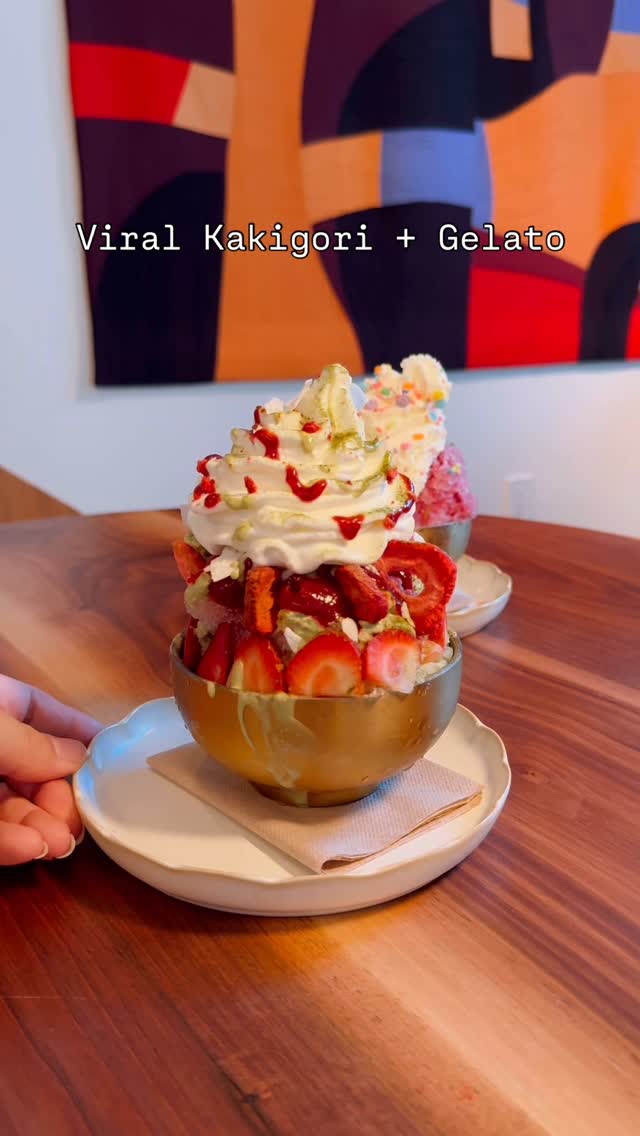 The cover image showcases a close-up of a bowl of vibrant Japanese Kakigori, a shaved ice dessert, topped with whipped cream, strawberries, and a drizzle of red sauce. There are other similarly arranged desserts in the background, suggesting multiple offerings. The setting is a wooden table, and a hand is seen holding the plate with the dessert bowl. Above the dessert, the text "Viral Kakigori + Gelato" is visible, written in a sans-serif font against a backdrop suggesting an artful interior or wall.