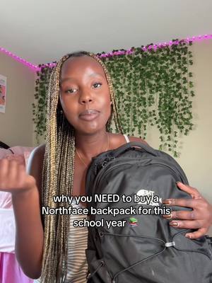 A young woman with long braided hair is holding a black backpack and pointing to her left with her other hand. She is in front of a wall covered in green vines and pink string lights, with a light color wall with a pink bed.