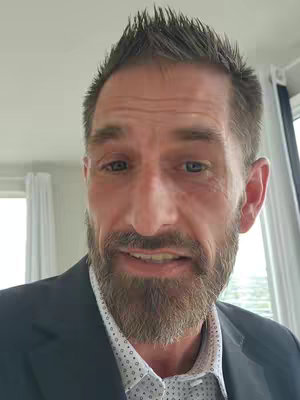 A close-up shot of a man's face, slightly tilted. He has short-spiked brown hair, a trimmed beard, and is wearing a dark suit over a white shirt with a dotted pattern. The background is out of focus, suggesting an indoor setting.