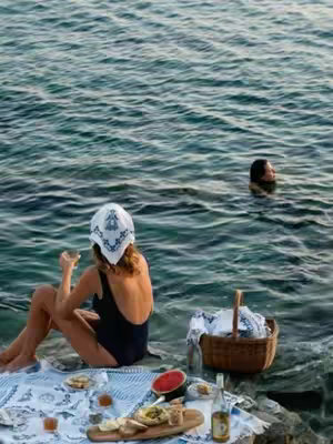 A woman wearing a white hat and a black swimsuit sits on a rocky shore with a picnic spread before her. The ocean stretches out in the background with another person swimming in the distance. The scene is brightly lit with colors that suggest a sunny day.