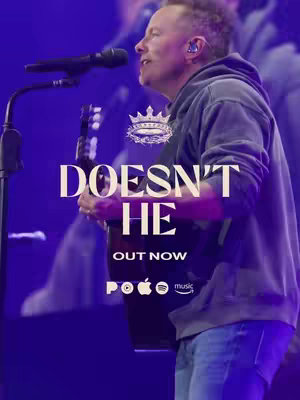The image is a vertical composition featuring a man singing into a microphone, with text overlay. The man, positioned slightly off-center to the left, is wearing a gray hoodie and jeans, holding a guitar. Behind him, a blurred background suggests a stage with purple lighting. In the foreground, large white text reads "DOESN'T HE" and smaller text "OUT NOW" is positioned below. Logos for music platforms (POCO, Apple Music and Spotify) are at the bottom. The overall composition is well-lit with a focus on the man and the title text.