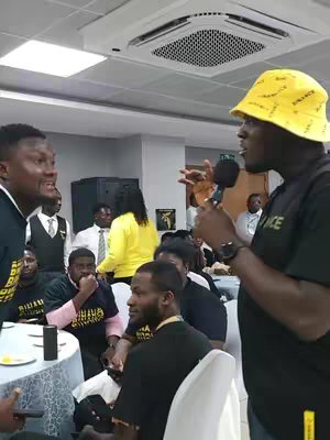 A group of people gathered in what appears to be a conference setting, with several men standing and speaking into a microphone. The man in the foreground wearing a yellow bucket hat with printed text and a black t-shirt. The background is filled with additional people, a suspended ceiling with air conditioning units, and event decor.