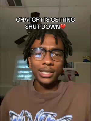 A young Black man with glasses is looking directly at the camera. The background is slightly blurred, showing a ceiling fan and some items on a shelf.