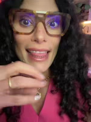 A close-up shot of a woman with dark curly hair wearing large, tortoise-shell glasses and a bright pink top. She is looking towards the camera with a slightly open mouth and her tongue touching her lip, suggestive of a playful gesture. Her hand holds a necklace pendant in the foreground. The background is somewhat blurred, suggesting a casual, indoor setting. 