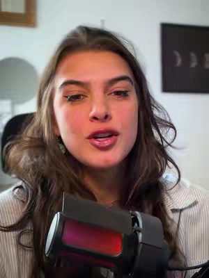A woman with long brown hair is speaking into a microphone. She has a light complexion and is wearing a striped button-down shirt. The background includes a glimpse of a framed picture, a circular object, and a plain wall.